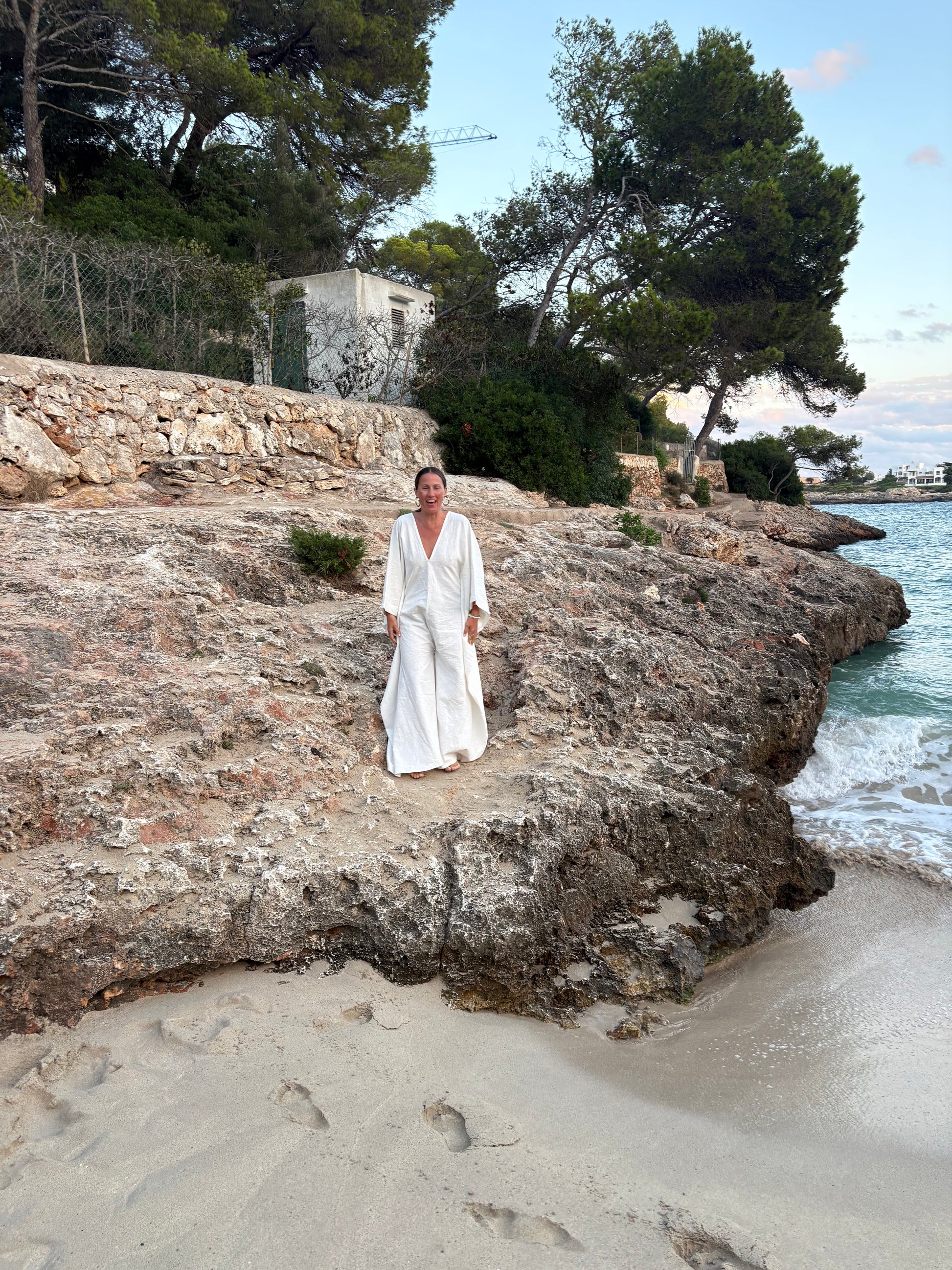A woman wearing a white long-sleeve maxi dress stands on a rocky beach next to the sea, with sand, trees, and a stone wall in the background.