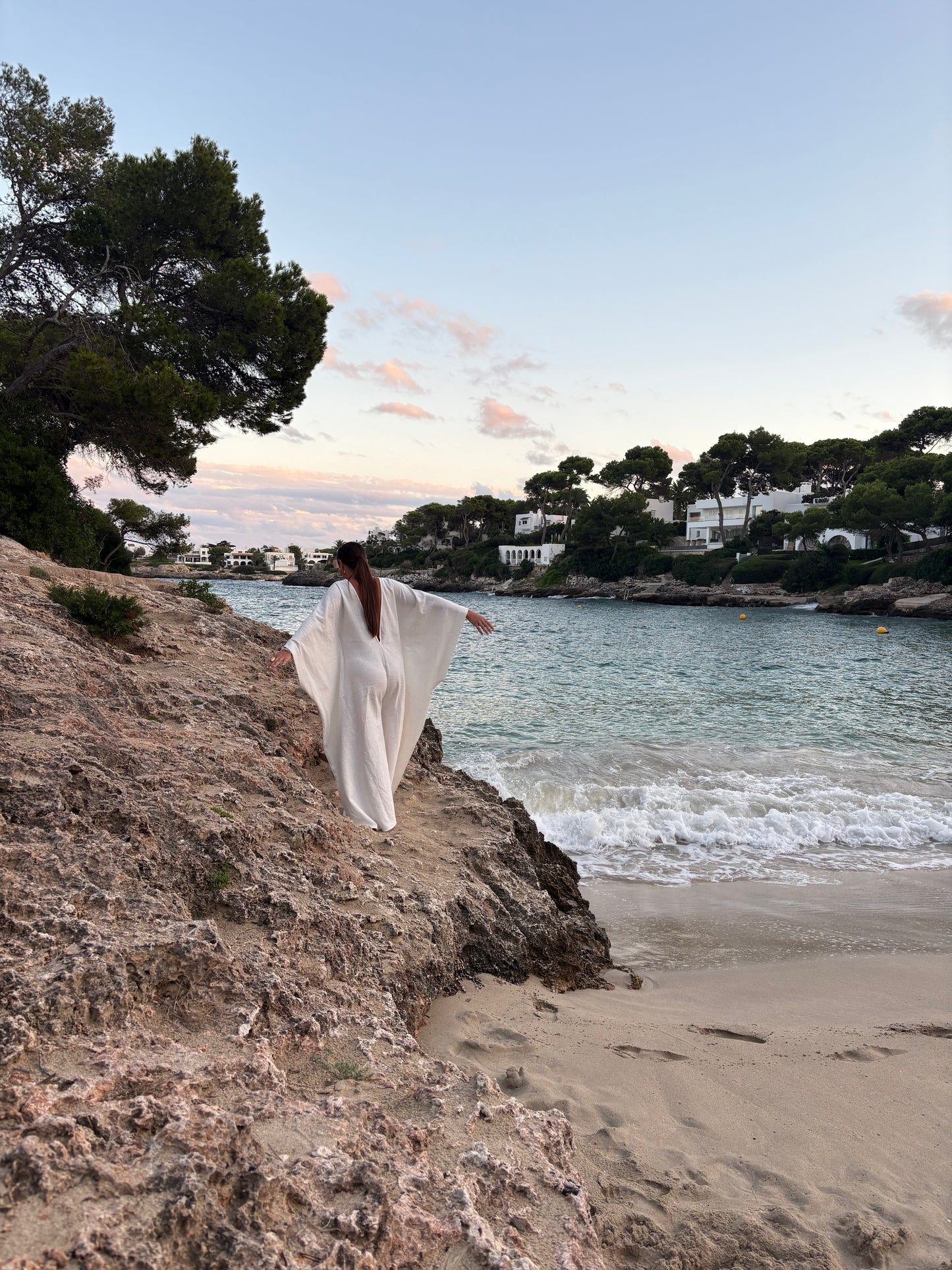 A woman in a flowing white maxi dress stands on a rocky beach cliff overlooking a calm bay with gentle waves, sandy shore, trees, and distant houses under a soft sky.