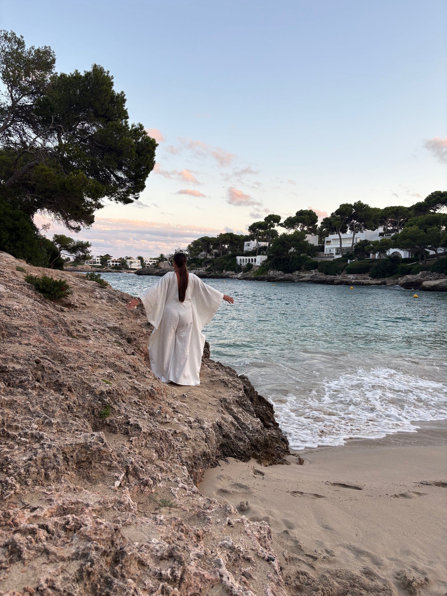 A woman in a flowing white dress stands on a rocky beach overlooking a calm bay with turquoise water, sandy shore, trees, and white buildings in the distance under a soft blue sky.