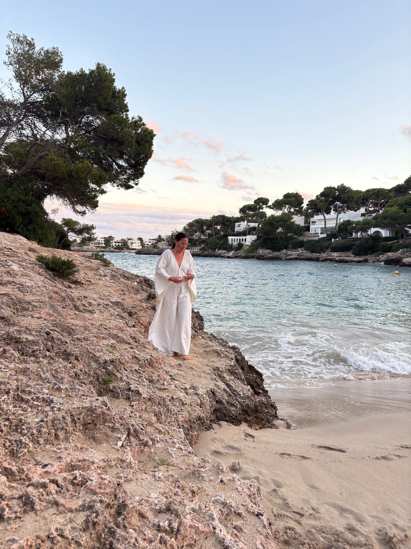 A woman in a flowing white robe stands on a rocky beach next to the sea, with sand, trees, and houses in the background under a soft sky.